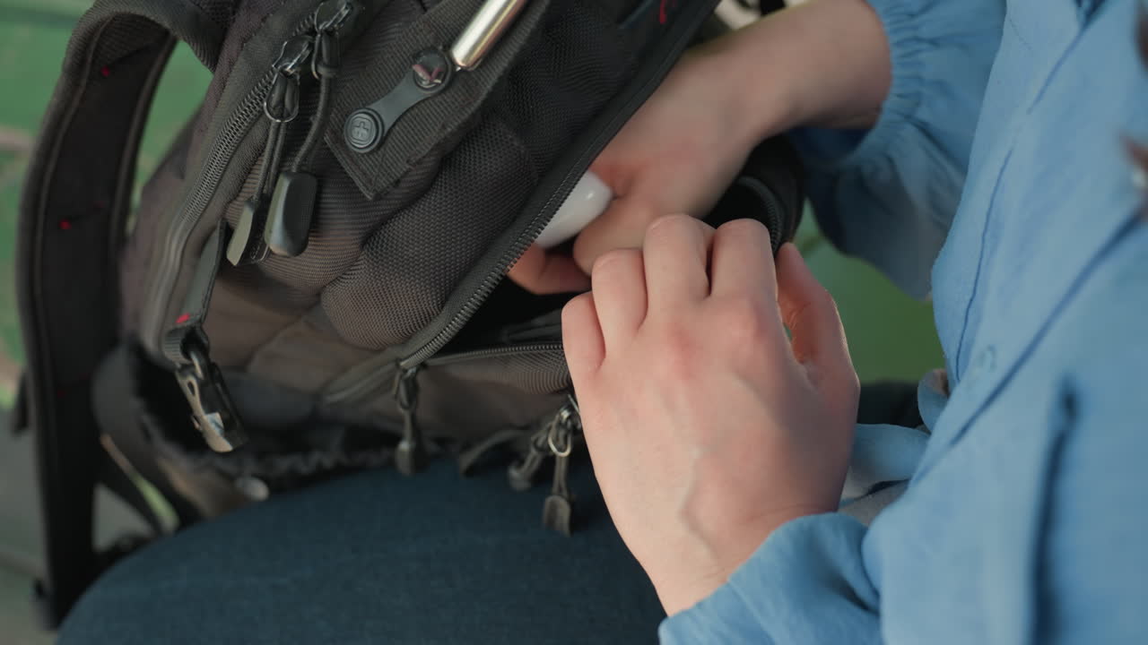 Primer plano de unas manos abriendo el bolsillo de una mochila y sacando un objeto pequeño, escena informal en un banco callejero, detalles de una cremallera y una manga azul claro, movimiento cuidadoso y concentrado que sugiere un viaje o una rutina diaria