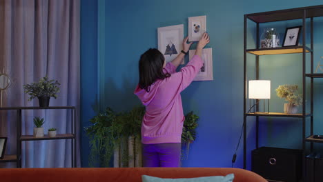 Mujer-Joven-Feliz-Poniendo-Una-Imagen-Moderna-Enmarcada-En-Una-Pared-Azul,-Decorando-La-Sala-De-Estar-En-Una-Casa-Nueva