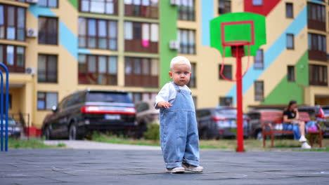 beautiful blond baby boy wearing loose jeans romper stands outdoors. kid looks focused at camera stepping back slowly. low angle view.
