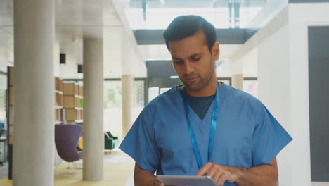 Male-Doctor-In-Scrubs-Using-Digital-Tablet-Walking-Through-Busy-Hospital-Building