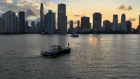 Toma-Aérea-De-Un-Hombre-En-Un-Yate-Cerca-De-La-Costa-De-Miami-Y-Paisaje-Urbano-Durante-La-Puesta-De-Sol