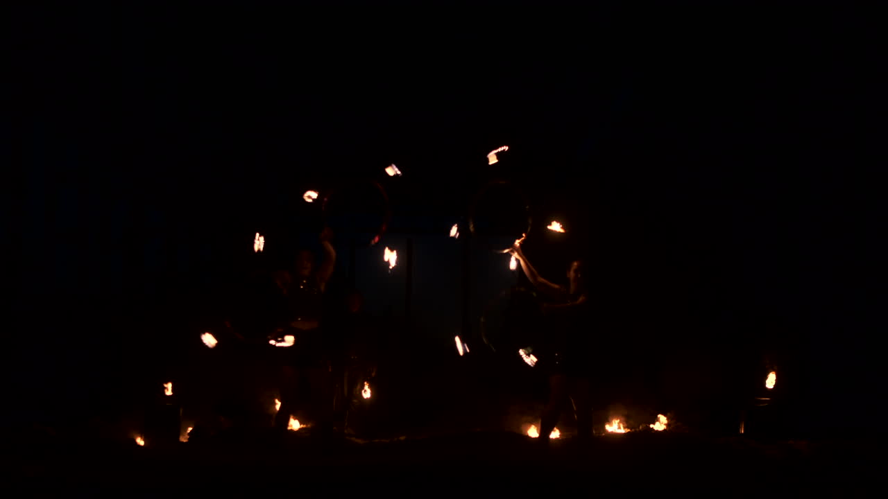 Three women with burning hoops dance with fiery torches in leather clothes in a dark hangar demonstrating a circus fire show in slow motion