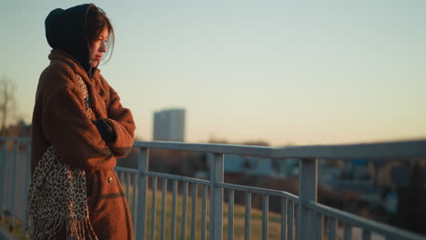 a melancholy girl stands close to a metal railing, wrapped in a warm brown coat and hoodie. she gazes into the distance, lost in thought