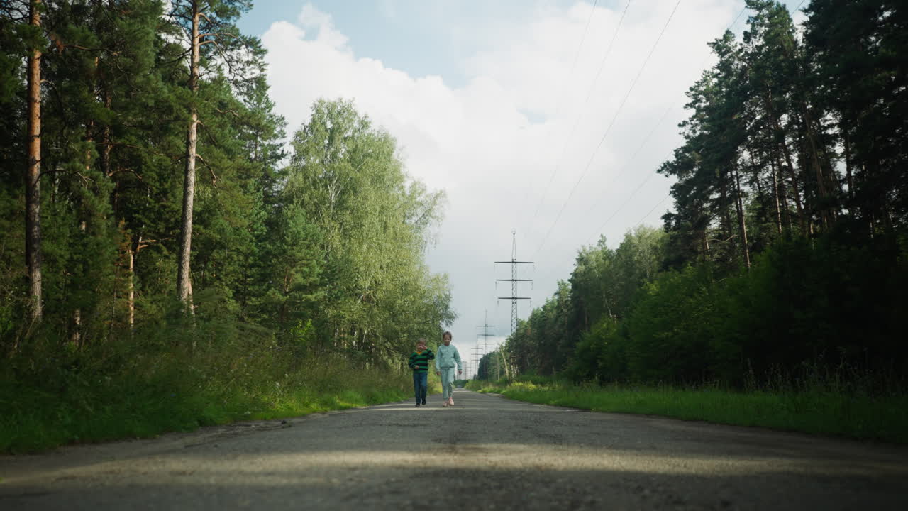 Tender moment as girl gently touches younger brother ear while walking together on quiet forest path under open sky, surrounded by lush greenery and tall pine trees on both sides