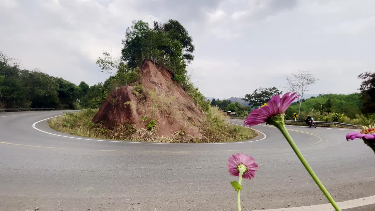 Motorcyclist rides past red cliffside curve on rural Thai road under bright overcast daylight