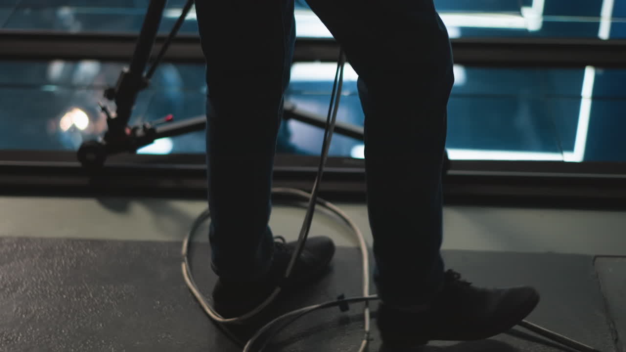 Foot of studio camera operator walking past camera dolly on track, floor with reflections visible, modern broadcast studio with lights, equipment, and glass reflections in background