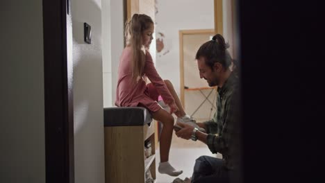 Shot-from-the-side-of-a-brunette-man-in-a-checkered-green-shirt.-A-lonely-father-helps-his-little-daughter-in-a-pink-dress-put-on-white-shoes-before-starting-their-walk.-A-brunette-man-helps-his-daughter-put-on-white-shoes-in-the-hallway-of-a-modern-apartment