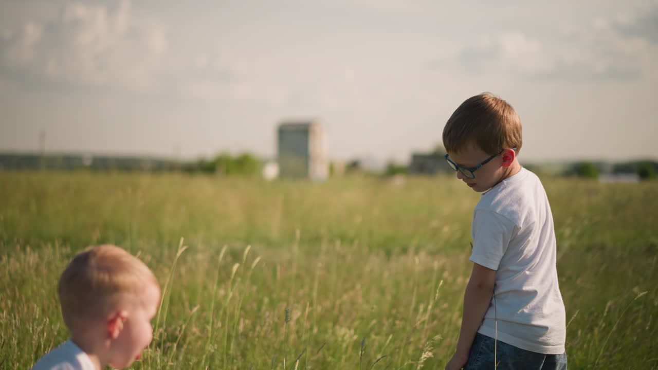 A back shot of a young boy walking through a grassy field, gently touching the tall grass. His younger brother joyfully jumps in to join him, and they exchange smiles, enjoying the outdoors together