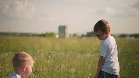 a back shot of a young boy walking through a grassy field, gently touching the tall grass. his younger brother joyfully jumps in to join him, and they exchange smiles, enjoying the outdoors together