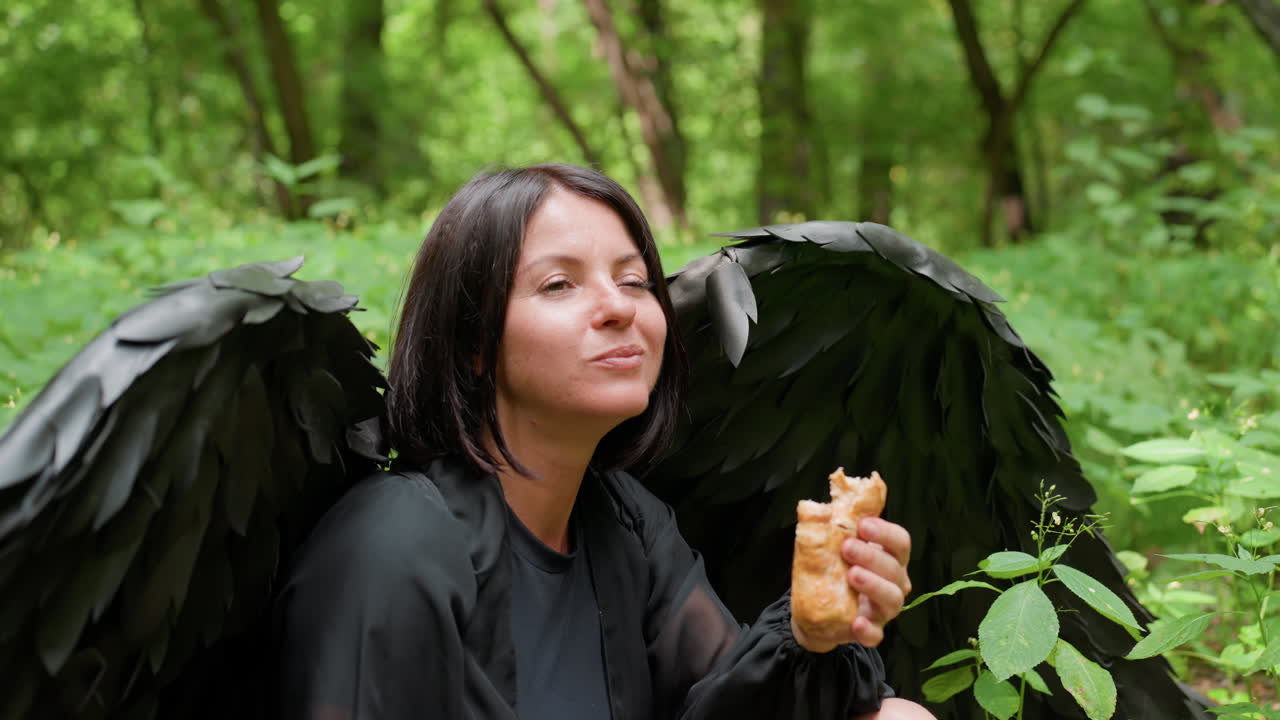 Young girl dressed in black Halloween costume with dark wings sits on green forest floor savoring burger, surrounded by lush plants and trees, creating calm mysterious atmosphere blending fantasy