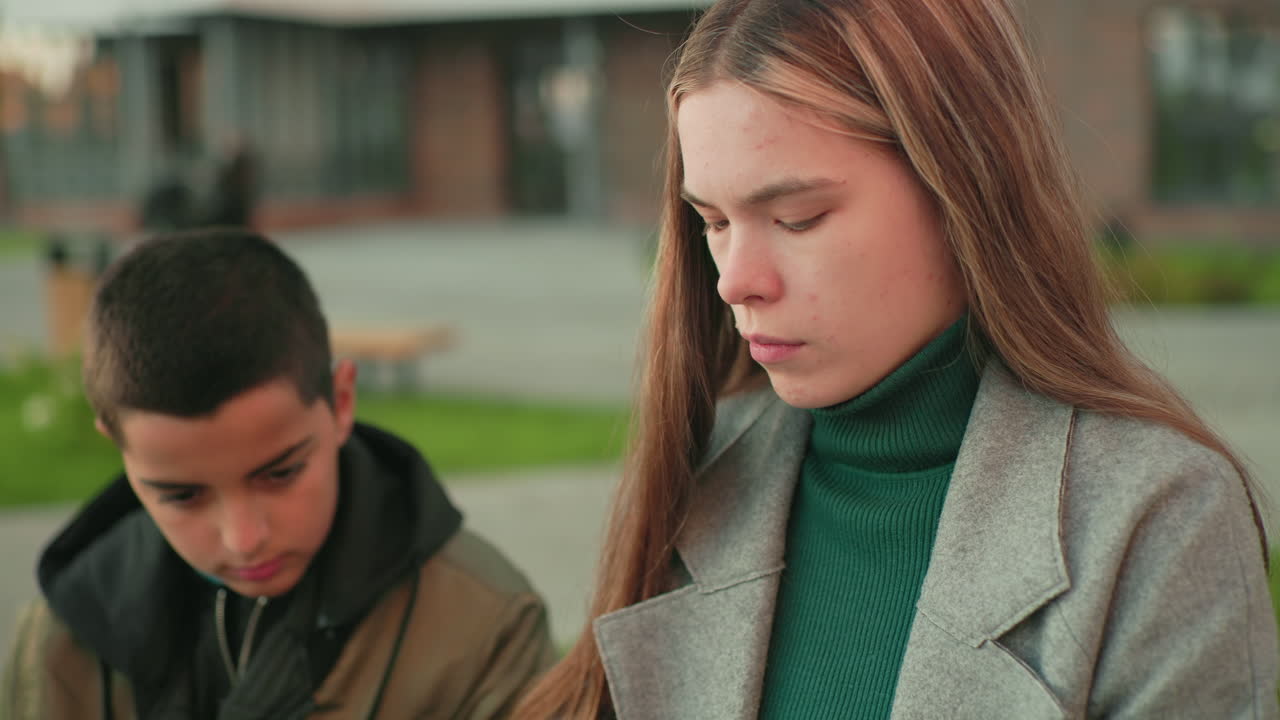 Close up of boy watching sister intently as she focuses on her task, both seated outdoors with soft blurred background, highlighting concentration