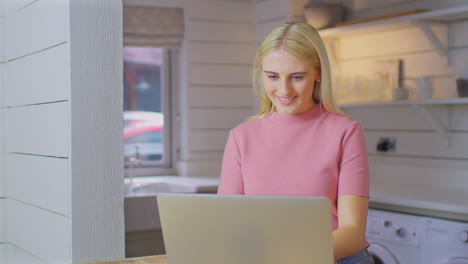 mujer joven de pie en el mostrador de la cocina con una computadora portátil trabajando desde casa