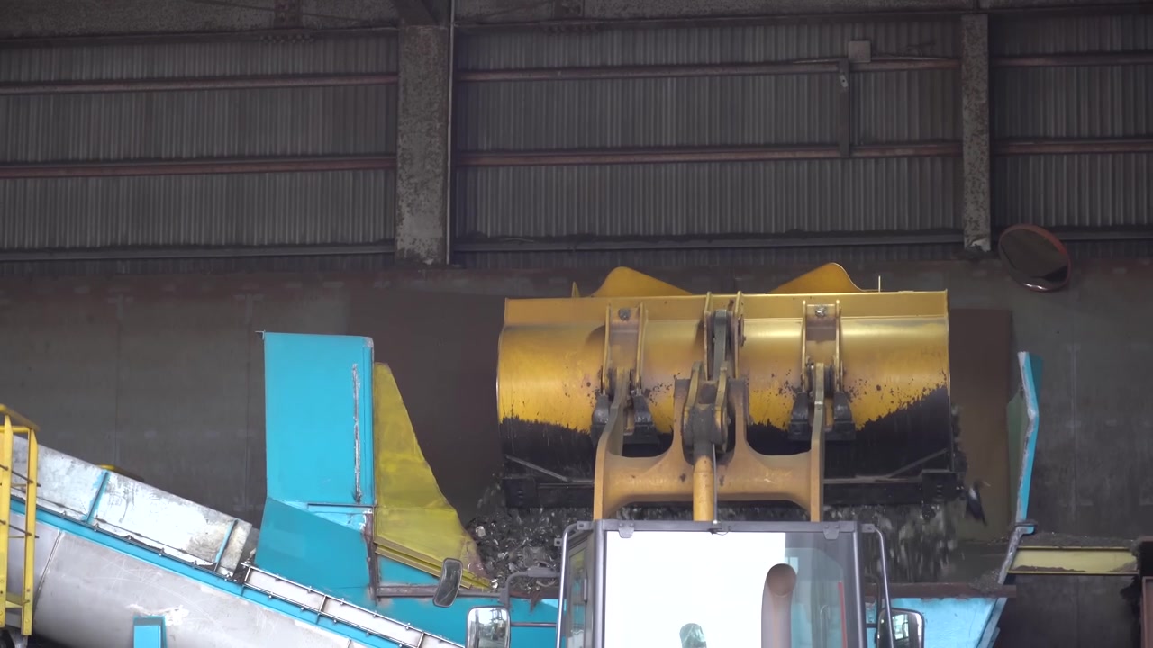 Yellow loader bucket feeding scrap material onto a blue industrial conveyor inside a recycling plant hall