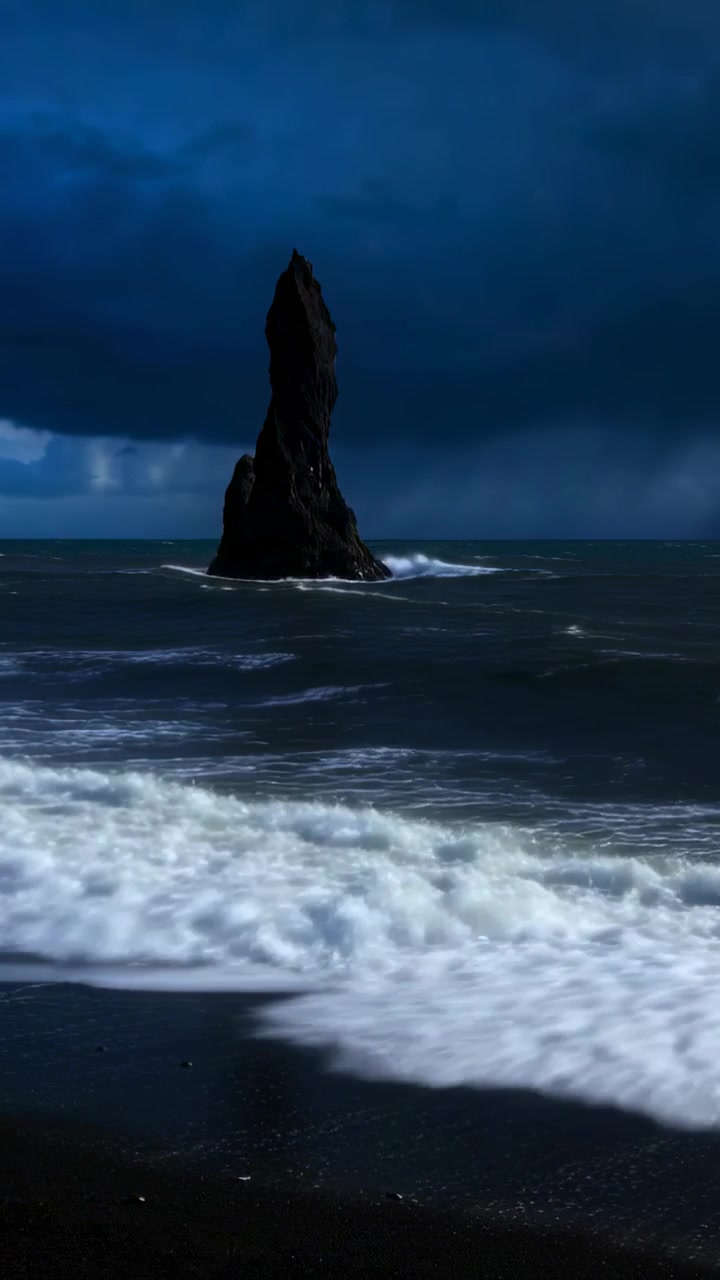 Vertical video: Breaking waves pushing sea stack on black sand shore, horizon swells driving foam