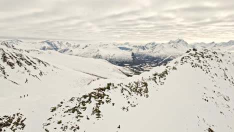 Group-of-people-on-edge-of-massive-snowy-mountain,-ready-to-ski,-aerial-view