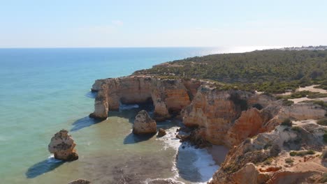 Rocky-ocean-spires-cast-long-shadows-from-cliffs-and-rough-waves-at-praia-da-marinha,-algarve,-drone