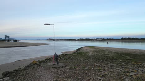 Solitary-lamppost-and-figure-amongst-rubble-on-river-shoreline-at-dusk-on-the-River-Wyre-estuary-Fleetwood-Lancashire-UK