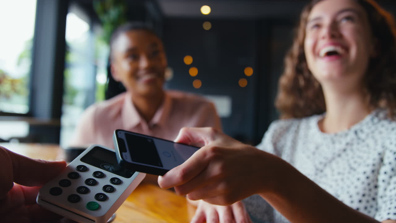 Woman In Coffee Shop Paying Bill With Contactless Mobile Phone Payment