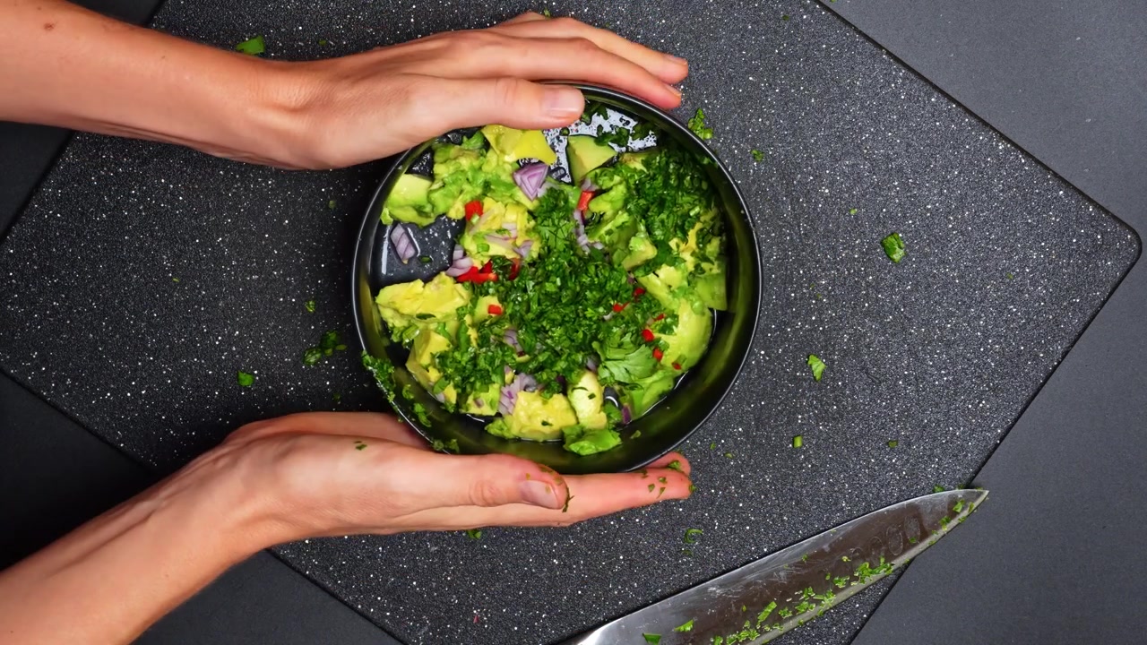 Mixing fresh ingredients to make guacamole in a kitchen. Hands are preparing guacamole by mixing avocados, onions, and herbs in a bowl on a kitchen counter.