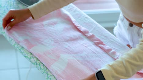 a woman arranges and irons a pink shirt on an ironing board in a brightly lit room, using careful hand movements and a steam iron
