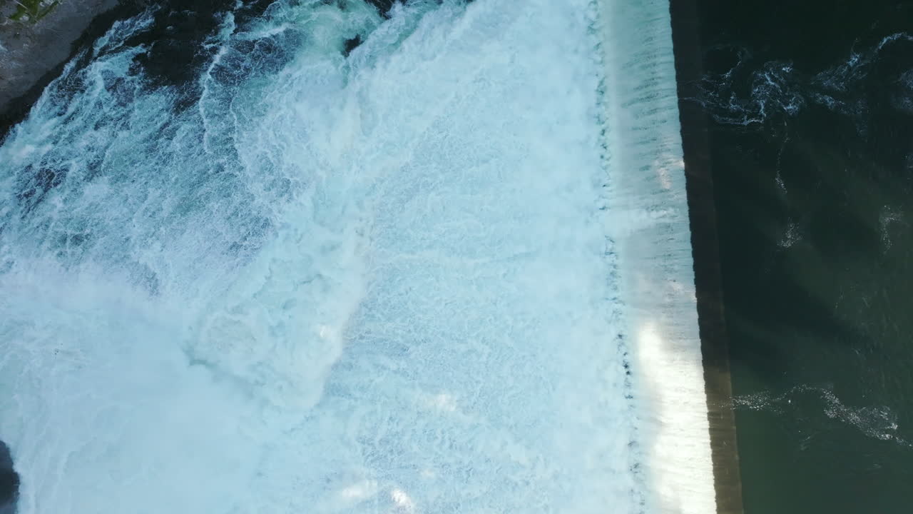 Water spills over a wide dam in a rush of white foam. The top-down view shows the full force of the river
