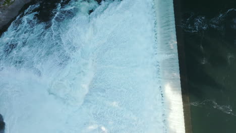 water spills over a wide dam in a rush of white foam. the top-down view shows the full force of the river