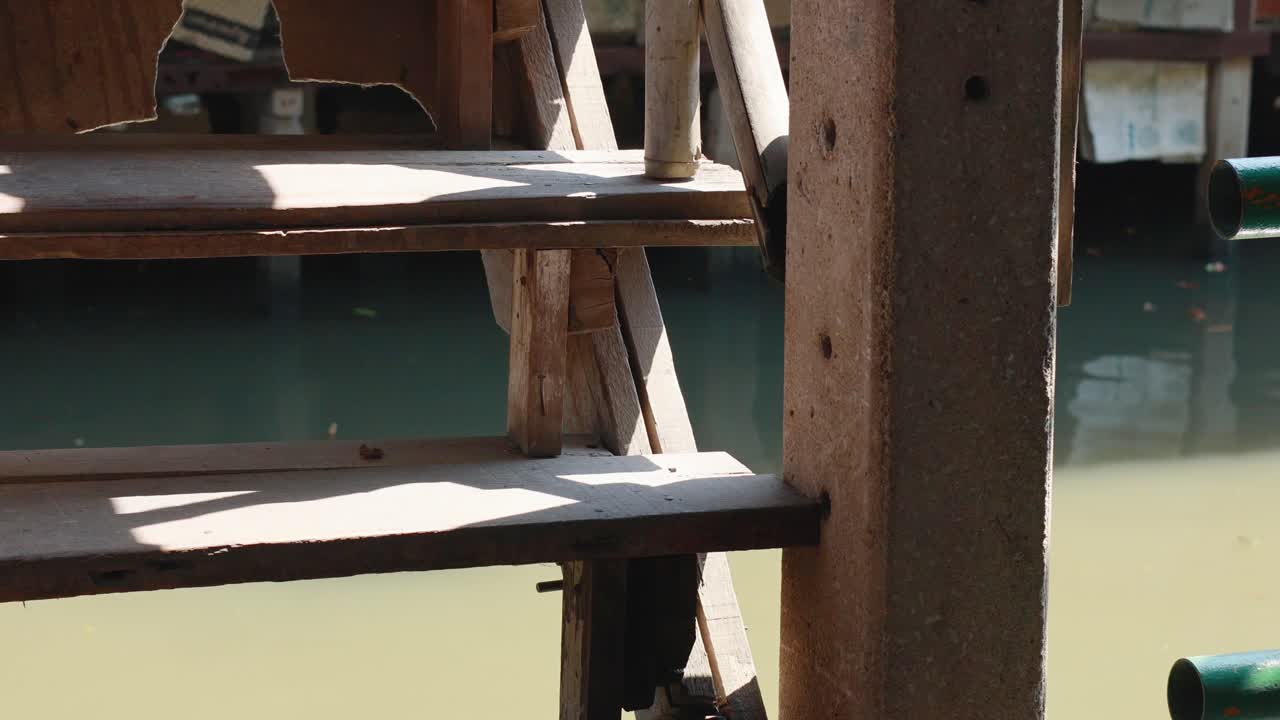 Wooden stairs with bamboo railing by a canal, capturing sunlight and shadows in a bustling Bangkok market setting