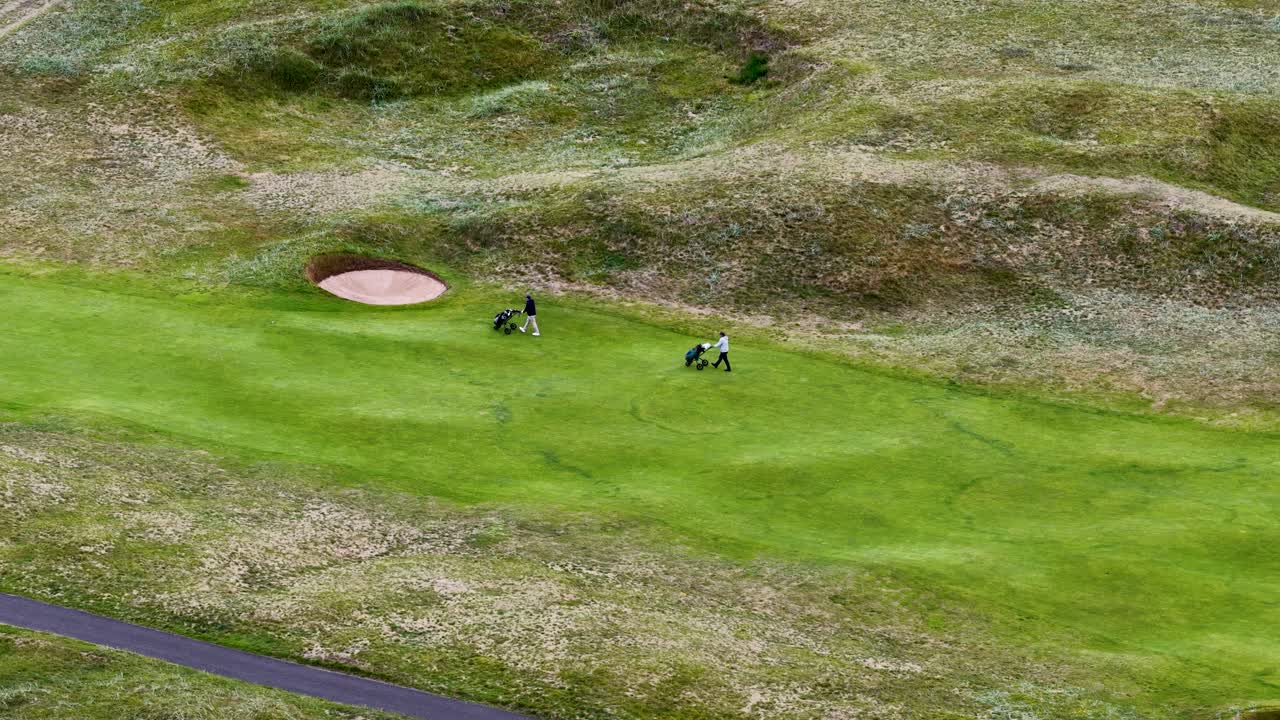 Two golfers walk with bags on lush fairway, drone camera gliding smoothly above, daylight