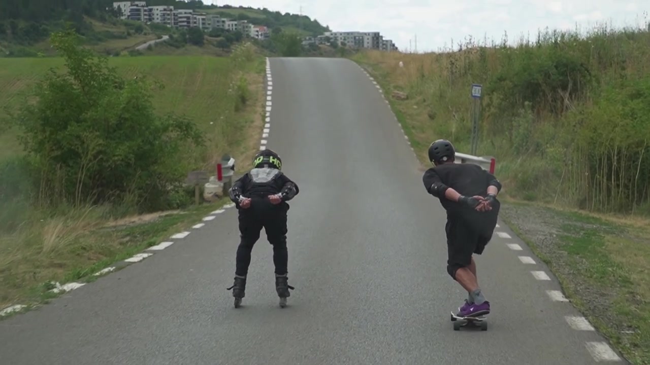 To personer, den ene på rulleskøjter og den anden på skateboard, suser ned ad bakke på en stille landevej.