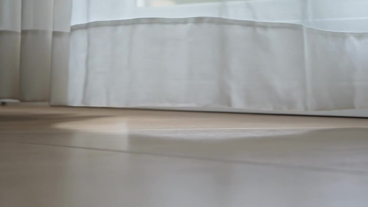 Shifting sun patch moving across wooden floor via window in home altering sheer curtain hem folds