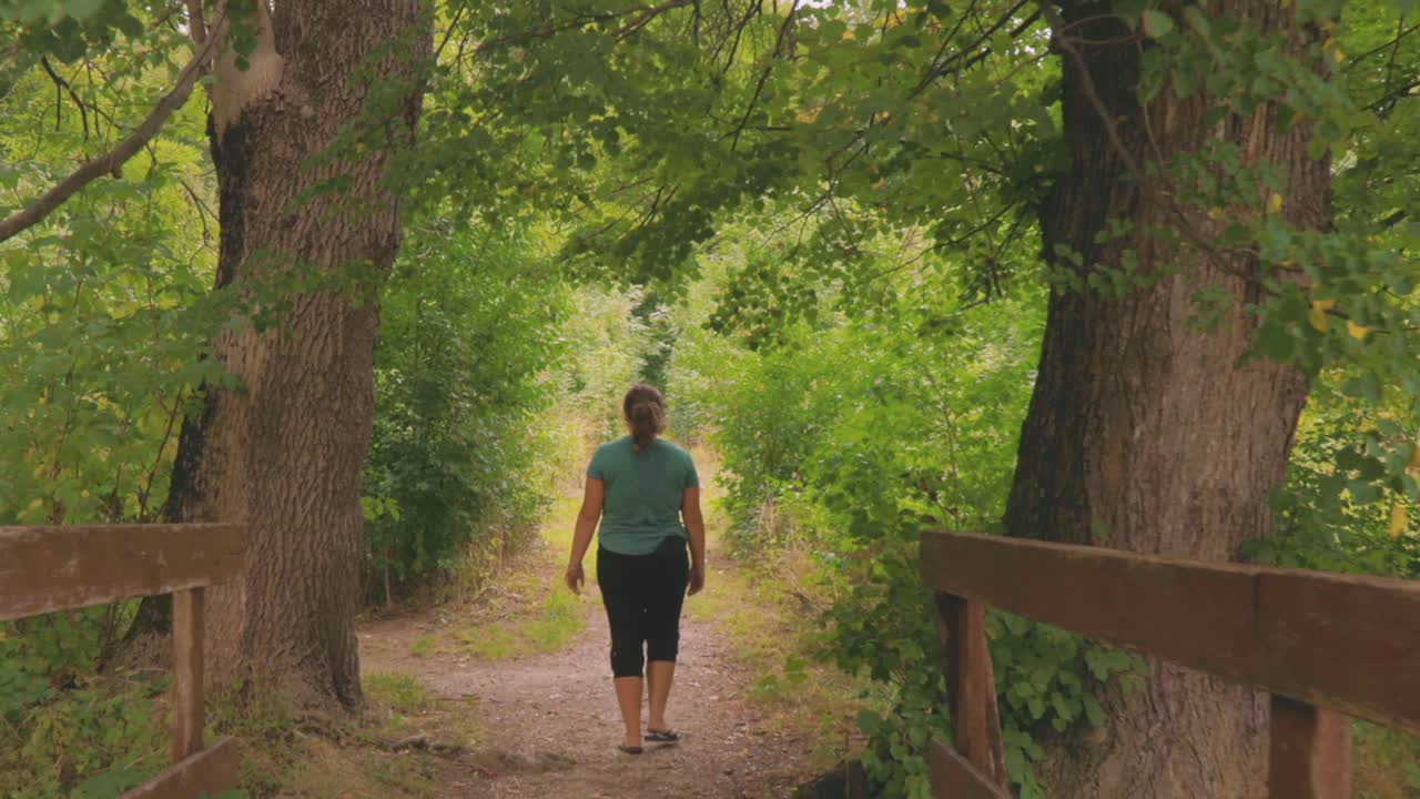 삼림 공원에서 나무 인도교를 걷고 있는 백인 여성을 따라