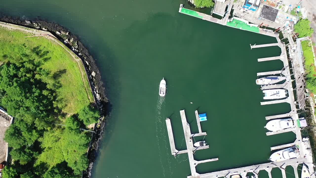An aerial view over a Brooklyn dock on a sunny day. The drone camera looks straight down and descends over a small boat in motion. The water is calm and green as the sun reflects on it