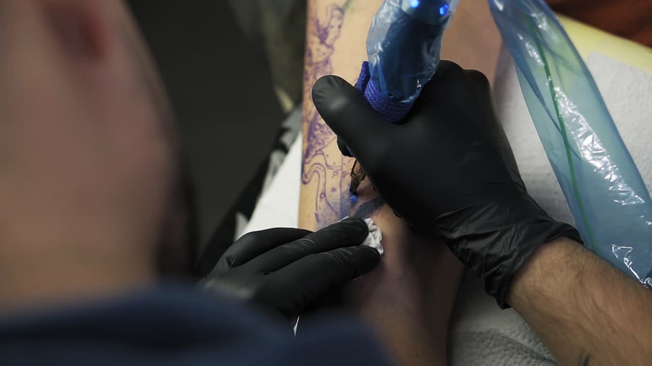 A close-up of a tattoo artist holding tattooing gun and makes a tattoo on a woman's arm using blue paint. Professional tattooist works in studio. High angle footage from the shoulder of master