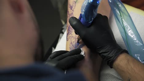 a close-up of a tattoo artist holding tattooing gun and makes a tattoo on a woman's arm using blue paint. professional tattooist works in studio. high angle footage from the shoulder of master