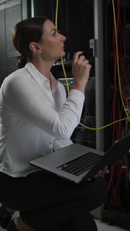 video vertical de una mujer caucásica usando una computadora portátil trabajando en una sala de servidores de computadoras