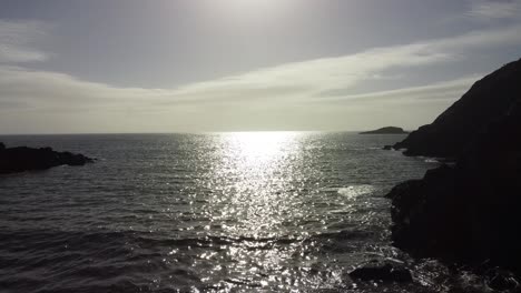 Aerial-View-Over-Calm-Waves-on-Rocky-Beach-with-Small-Island-and-Bright-Sun