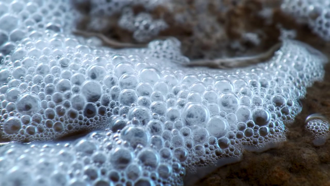 Densa striscia di schiuma di sapone e bolle fittamente raggruppate che poggiano e si espandono lungo una superficie scura e porosa, mostrata in un'inquadratura panoramica ravvicinata.