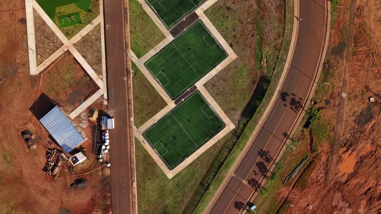 New five-a-side football soccer fields sit alongside a curved urban road and construction site, bright green synthetic pitches bordered by walkways, in Posadas, Misiones, Argentina