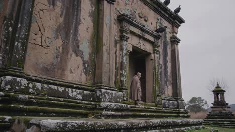 monk descending steps of ancient stone temple with weathered facade and birds perched on roof, evoking spirituality, history, and nature's presence