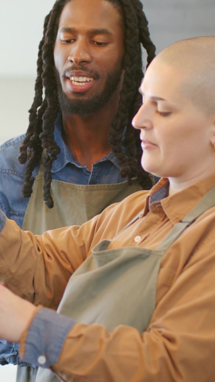 Vertical video: Leaning man teaching woman at counter, handling utensil in aprons and collared tops