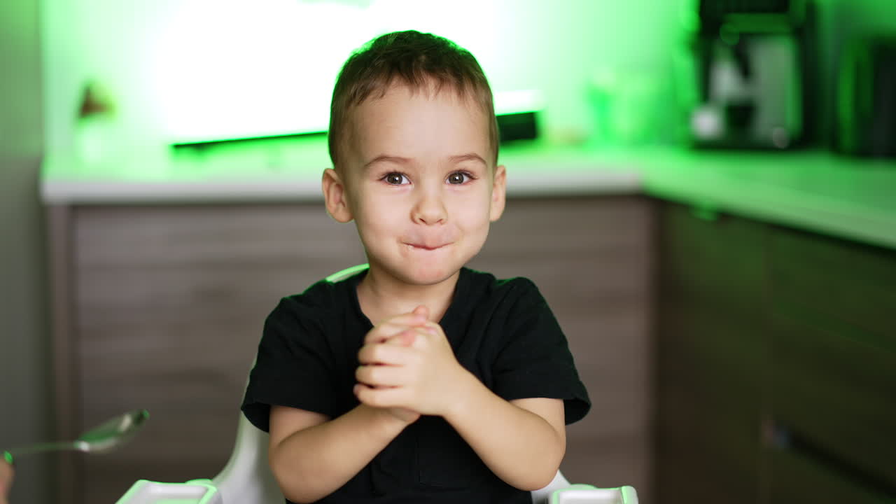 Feeding a cute Caucasian toddler boy from spoon. Smiling child chews his food and plays with hands. Blurred backdrop.