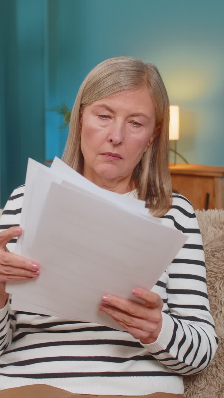 Mature woman reviewing documents and bills on sofa expressing uncertainty and frustration at home
