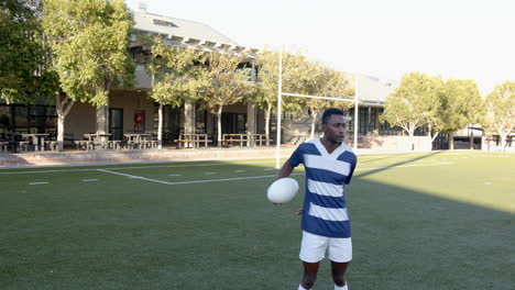 holding rugby ball, african american man standing on field in front of school building, copy space