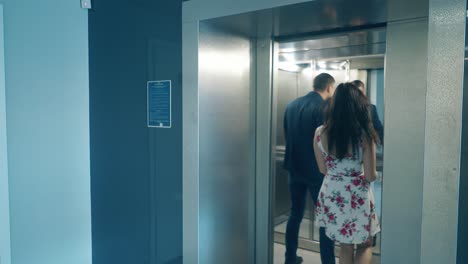 un hombre de traje entra en un ascensor gris sosteniendo a una chica morena