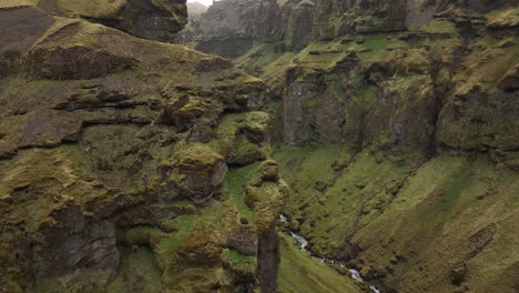 aerial shot of steep, moss-covered cliffs towering over a narrow canyon with a river flowing through the rugged landscape of brókarjökull, iceland