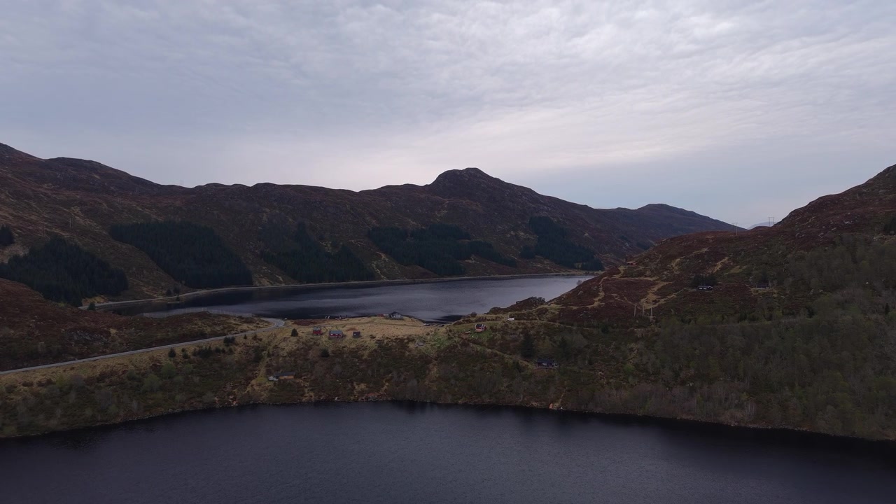 En stille norsk dal med mørkt vand, der reflekterer den overskyede himmel. Barske bakker og spredte skove skaber et fredfyldt landskab. Velegnet til rejseindhold eller naturdokumentarer.