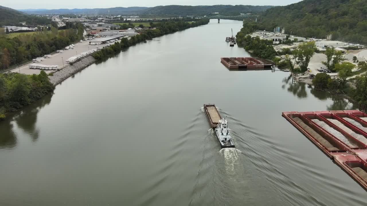 Aerial drone footage of towboat guiding cargo barge along calm Kanawha River surrounded by forested hills, industrial docks, and quiet roads in scenic West Virginia, United States