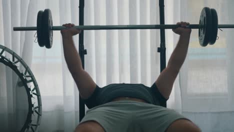 man doing bench press workout with a barbell in the gym by a window with a trampoline