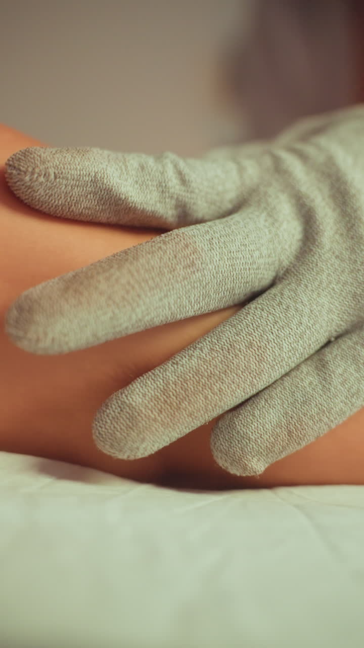 mani guantate eseguono un massaggio microcorrente alle gambe, guanti conduttivi testurizzati scivolano su polpaccio e piede fornendo una stimolazione sottile, primo piano in un ambiente clinico con illuminazione calma, terapista concentrato