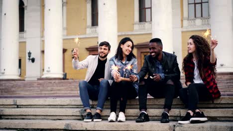 cinemagraph loop - happy young people friends in casual clothes sitting on stairs outdoors with burning bengal lights and smiling. youth, happiness and city concept.